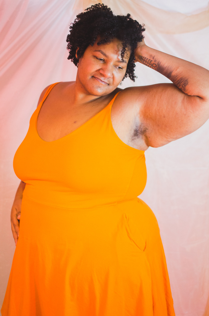 Kumi, eine Schwarze, dicke Person, in einem orangefarbenen Kleid, greift ins eigene ohrlange lockige Haar, so dass die Achselhaare sichtbar sind. Kumi schaut lächelnd nach unten.
