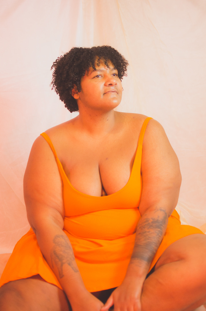 Kumi sitzt auf dem Boden mit einem Bein seitlich angewinkelt und den Händen am Unterschenkel in einem kurzen orangenen Kleid mit Spaghettiträgern. Kumi schaut leicht lächelnd nach rechts. Kumi ist eine Schwarze, dicke Person mit halblangem, lockigen Haar, sehr dicken Oberschenkeln & Oberarmen und sichtbaren Tattoos an den Unterarmen.